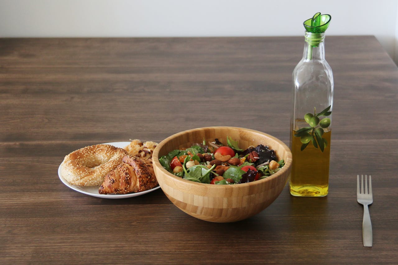 A vibrant vegetable salad with bagel and croissant, olive oil on a wooden table.