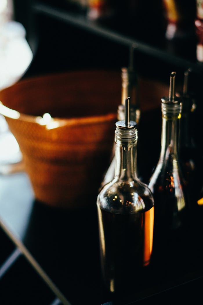 Close-up shot of clear glass bottles filled with liquid, likely olive oil, on a dark counter beside a wicker basket.